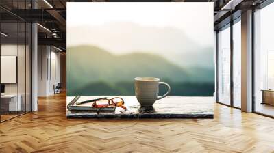  A coffee cup, eyeglasses and a note book on wooden table with sunrise and mountain  bokeh background. A start of new day with hot beverage. Business concept. Wall mural