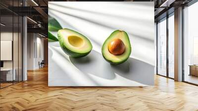 Two halves of an avocado with a single pit, illuminated by sunlight on a white surface with a leafy green stem Wall mural