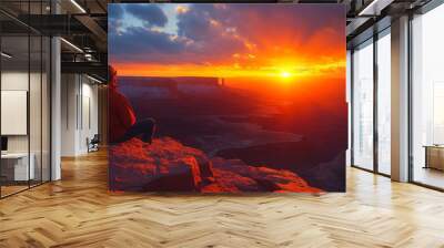 Person sits on a rocky cliff, observing a vibrant sunset illuminating a vast canyon landscape with rays of sunlight Wall mural