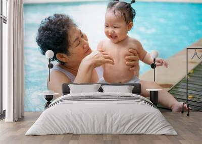 Senior asian woman is enjoying time with her adorable one year old grandchild in a swimming pool. Wall mural