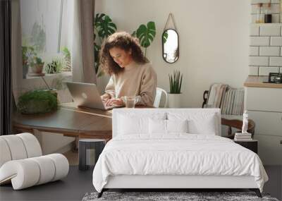 Charming young woman typing on laptop computer in a kitchen. Wall mural