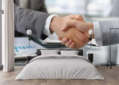 Close up of two business people shaking hands over computer desk with pen and paper on it. Wall mural