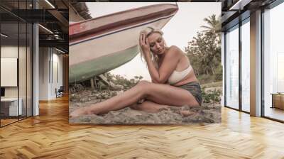 beach lifestyle portrait of young attractive and relaxed blond woman sitting on sand at tropical beach next to old stranded boat in exotic island holidays travel destination Wall mural