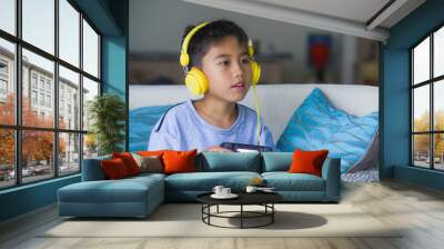 oung hispanic little kid excited and happy playing video game online with headphones holding controller enjoying having fun sitting on couch in child gaming addiction Wall mural