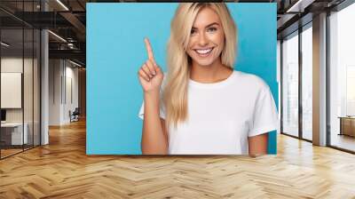 Blonde Caucasian girl in Hawaiian T-shirt stands against blue wall, smiling and gesturing number two with her hand, counting down. Wall mural