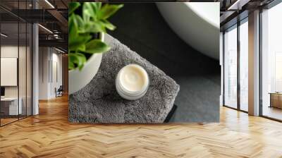 Men's facial cream in an open jar with a blank lid placed on dark grey folded towels alongside a green plant and a washbasin in a bathroom. Overhead perspective of cosmetic product packaging mockup. Wall mural