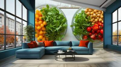 Two glass bowls filled with a variety of food items including green salad leaves, red and yellow cherry tomatoes, and chickpea salad, placed on a white wooden table. Wall mural