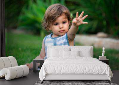 A 1.5 years old boy is sitting on a green grass in a garden and raising his hand while playing. Wall mural