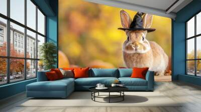 A cute rabbit wearing a black witch hat poses among pumpkins and autumn leaves, capturing a festive fall vibe. Wall mural