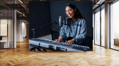 Happy cheerful pretty smiling of young Asian woman vocalist Wearing Headphones recording a song front of microphone and playing the keyboard during rehearsal of her band in a professional studio Wall mural