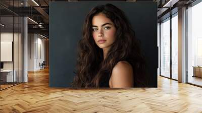 A beautiful woman with long, curly hair, wearing a black dress, poses for the camera against a white background Wall mural