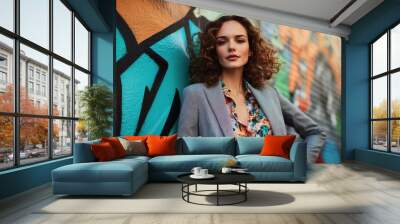 A confident woman in an elegant blazer and floral blouse stands against the backdrop of vibrant street art, her curly hair flowing gracefully as she poses for the camera with one hand on her hip Wall mural