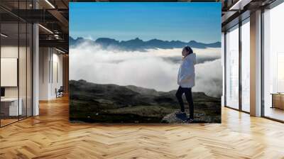 A young woman standing and enjoying the view of misty clouds covering the mountain range in front. in cold weather and has a beautiful view Wall mural