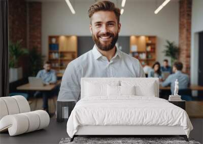 Smiling young bearded businessman in white shirt standing in light spacious office cafeteria and holding colorful folders while looking at camera Wall mural