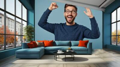 Young handsome man with beard wearing casual sweater and glasses over blue background very happy and excited doing winner gesture with arms raised, smiling and screaming for success Wall mural