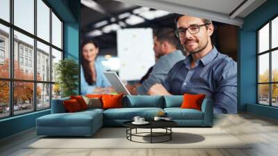 The creative process. Portrait of a man sitting at a table in an office using a digital tablet with colleagues working in the background. Wall mural