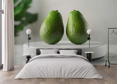 Two ripe avocados on a white pedestal, with a blurred green leaf in the background. Wall mural