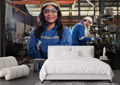 Portrait of professional Black female worker in protective safety uniform and helmet looking at camera, arms crossed and smiling, with engineers team behind her in a metalwork manufacturing factory. Wall mural