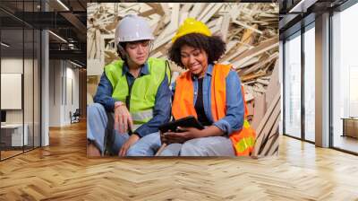 Two female workers and colleagues in safety uniforms and helmets, supervisor teams sit on pile of used paper for rest, talking and laughing in paper manufacture factory, recycling industry occupation. Wall mural