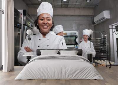 Young African American female chef in white cooking uniform looks at camera, arms crossed and cheerful smile with food professional occupation, commercial pastry culinary jobs in a restaurant kitchen. Wall mural