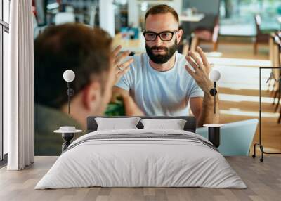 Attractive bearded man with glasses in a cafe working on laptop and chatting with a friend Wall mural