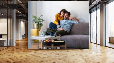 Couple using mobile phone at home stock photo. Positive couple is surfing internet on smart phone while sitting on sofa Wall mural