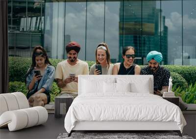 Group Of Five Happy Multi Ethnic Friends Using Mobile Phones Outdoors. 
Smiling friends watching or typing something on the phone, talking and laughing while sitting in a row on a bench in the city. Wall mural