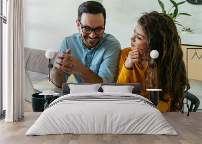 Happy Couple Paying Bills Together at Home. 
Smiling man and woman talking about family budget and savings while sitting at table with laptop computer, bills and two cups of coffee. Wall mural