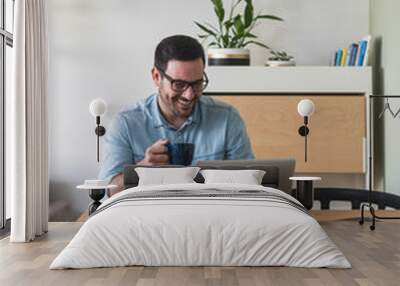 Happy smiling young man watching and working on computer laptop at home and drinking coffee or tea stock photo Wall mural