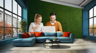 Two young people sitting over a green grass wall, using a laptop Wall mural