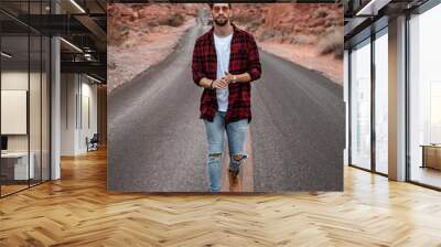 Man walking alone on the middle of the road, wearing plaid red shirt and sunglasses. Canyon road with red rocks are by the road. Wall mural