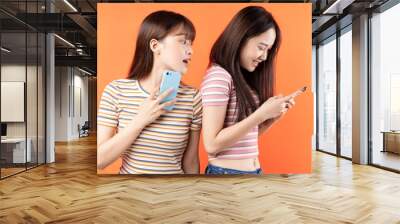 Two beautiful young asian girls are using mobile phones on orange background Wall mural