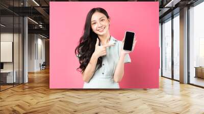 Young Asian woman holding phone in hand and pointing at it with playful expression Wall mural