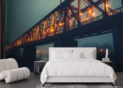 An iron railway bridge lit up in soft orange and yellow hues, stretching over a river, with trains passing in the distance. Industrial and dramatic Wall mural