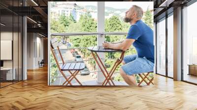 Bearded man sitting at wooden table on hotel balcony enjoys summer vacation. Male tourist explores sunny tropical resort town from room balcony Wall mural