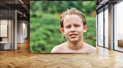 Cute schooler with wet blond hair stands against blurry lush trees and greenery in countryside. Portrait of relaxed boy closeup Wall mural