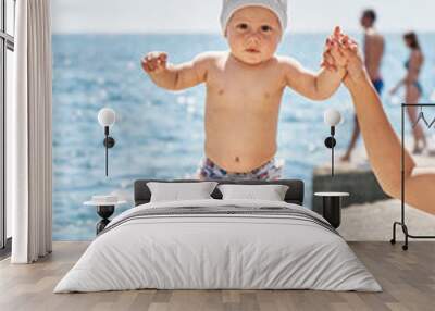 Infant baby tries to walk on stone pier with mother help. Boy enjoys walking and visiting for first time sunlit sea beach resort with family Wall mural