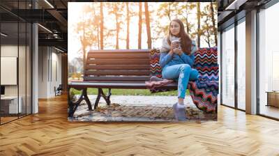 Young and attractive girl sitting on a bench in autumn park and drinking coffee Wall mural