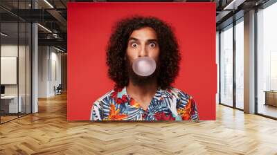 Amazed pretty brown-eyed bearded guy with dark curly hair making bubble with gum and looking surprisedly to camera, standing over red background in shirt with floral print Wall mural