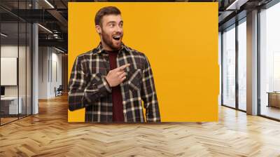 Astonished handsome young bearded man in checkered shirt with opened mouth looks surprised and pointing by finger away to the side over yellow background Wall mural