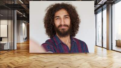 Attractive positive brunette curly man with beard holding camera and smiling sincerely while making selfie, wearing striped multi-colored shirt while posing over white background Wall mural