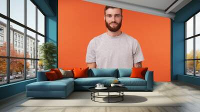 Attractive young bearded tattooed brunette man with trendy haircut standing over orange background, keeping hands along body and looking at camera with charming smile Wall mural