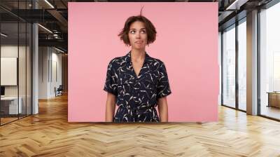 Attractive young brunette lady with short haircut wearing patterned elegant dress, posing over pink background with hands down, looking upwards and blowing on her hair Wall mural