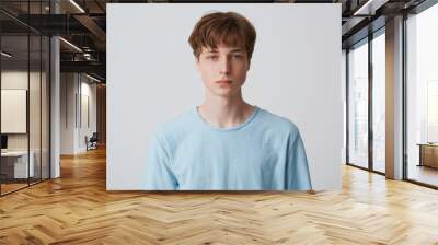 Close up face of a young man without emotions. Beautiful emotionless guy in a blue t-shirt looking to the camera, isolated over white background Wall mural