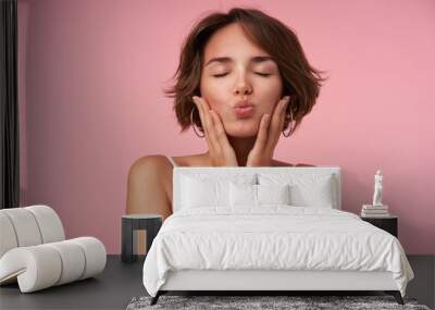 Close-up of pleasant looking brunette female with short haircut posing over pink background with eyes closed, holding her face with raised hands and folding lips in kiss Wall mural