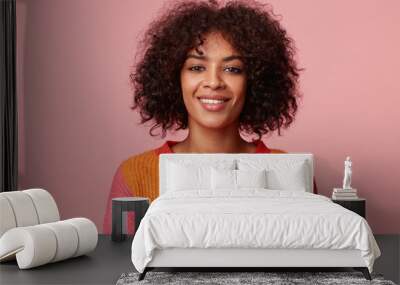 Close up portrait of positive optimistic charming african american girl with afro hairstyle looks with pleasure, with friendly smile, wearing colorful longsleeve, isolated on pink background Wall mural