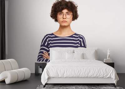 Closeup of calm serious emotionless young curly brownhair woman wears white and blue stripped sweatshirt and transparent black-framed round glasses keeps arms crossed isolated over white background Wall mural