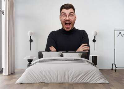 Excited young lovely bearded brunette male crossing arms on his chest and looking amazedly at camera with wide eyes and mouth opened, standing over white background Wall mural