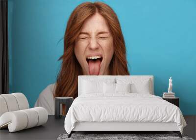 Funny shot ot young joyful redhead lady with natural makeup fooling while posing over blue background, frowning face and sticking out her tongue with closed eyes Wall mural