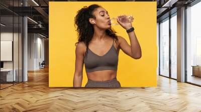 Happy beautiful african american young sportswoman listening to music using wireless earphones and drinking water from bottle isolated over yellow background Wall mural